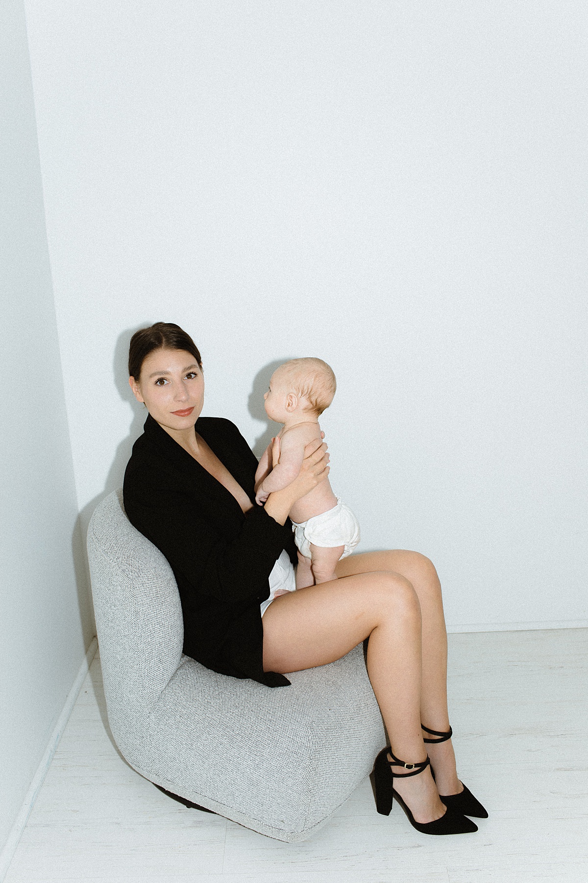 A motherhood photography session in a studio with white walls and floors. The mother wears a black blazer and heeled point shoes.
